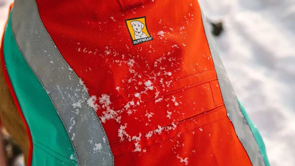Orangefarbener Hundemantel von Ruffwear mit grauen Reflexstreifen, ideal für winterliche Bedingungen im Schnee.