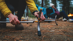 Person stellt Campingstuhl auf, im Hintergrund entspannt jemand am Lagerfeuer im blauen Stuhl.