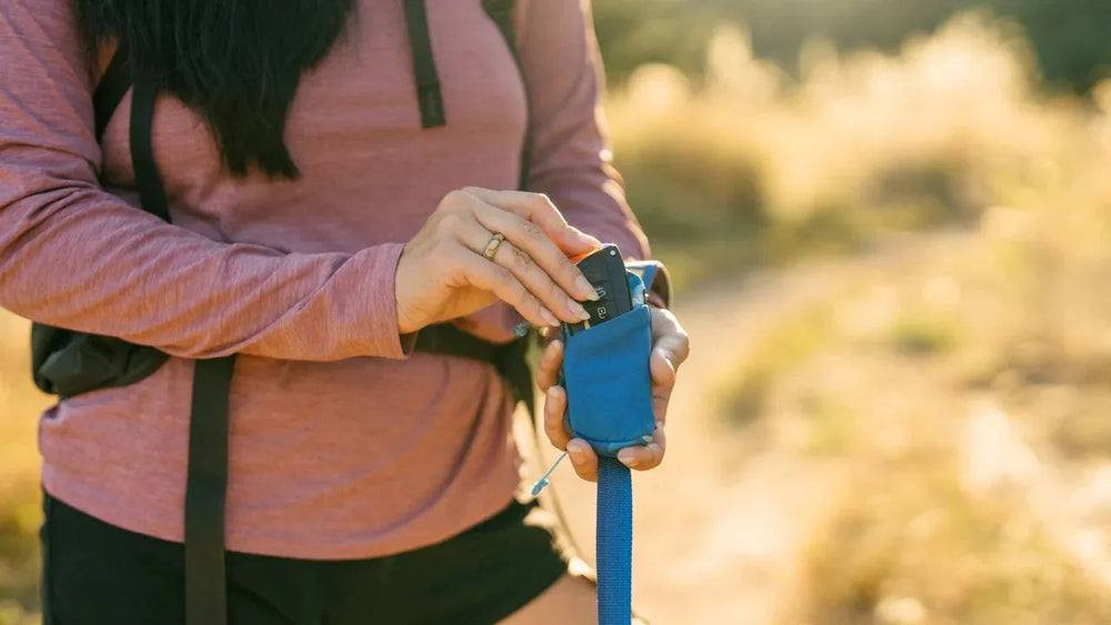 Person mit rosa Shirt steckt Kamera in blauen Stash Bag Mini™ Beutelspender während sie eine Sporttasche trägt.