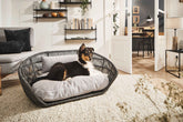 Modernes Hundebett TUDOR PRADO im geflochtenen Korbrand mit Hund im Wohnraum, graues Sofa und Bücherregal im Hintergrund.