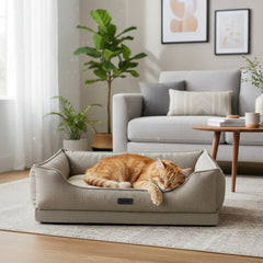 Gemütliche Katzenliege "CALBU" mit orangefarbener Katze in modernem Wohnraum neben grauem Sofa und Pflanze.