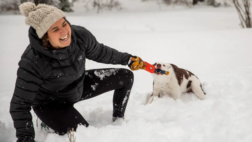 Frau im Schnee spielt mit ihrem Hund, der am Pacific Loop™ Spielzeug zieht – fröhliche Momente in Winterlandschaft.