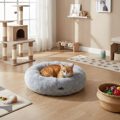 Gemütliches Komfortbett Donut "Nuru" für Katzen, grauer Plüsch, mit entspannter orangefarbener Katze im Hintergrund.