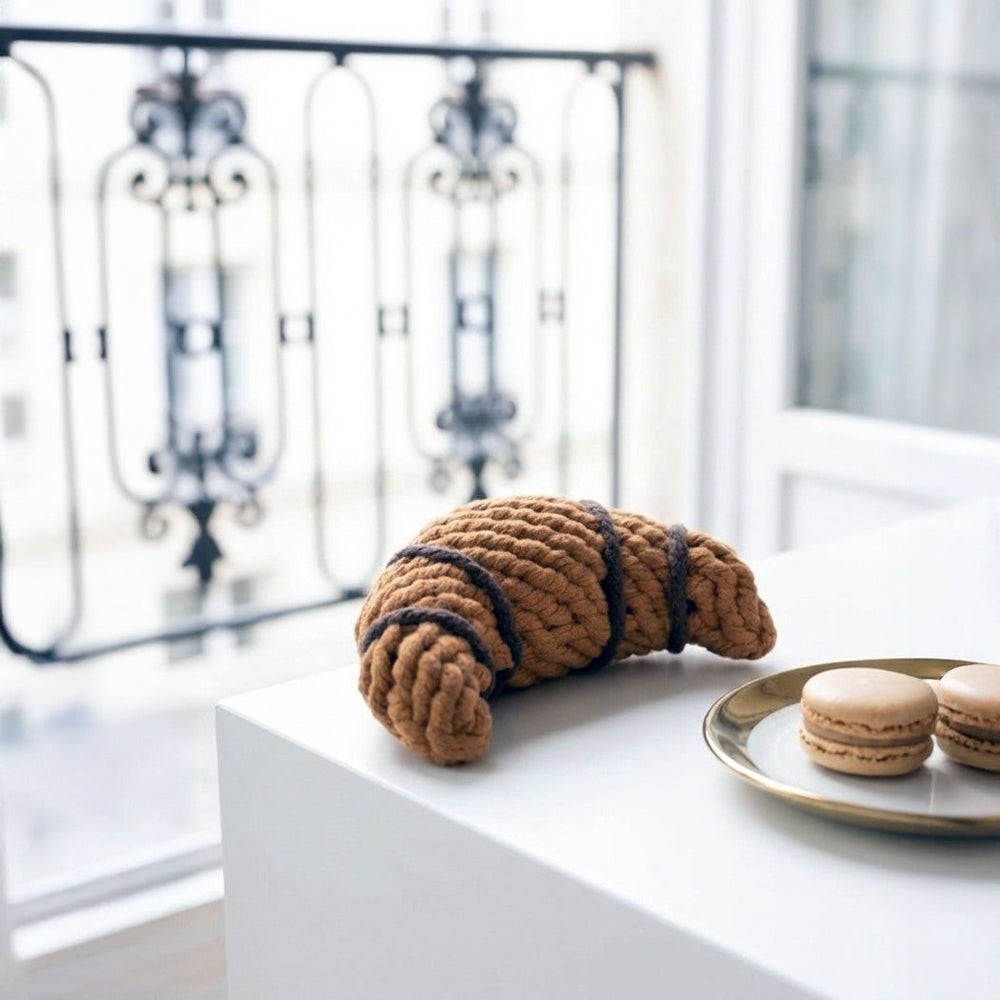 Geflochtenes Croissant aus Stoff auf weißem Tisch, begleitet von Macarons auf goldenem Teller. Hintergrund mit Fenster.