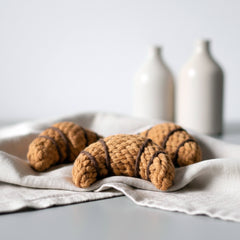 Handgemachte gehäkelte Croissants in Brauntönen auf hellem Tuch, dekoriert mit cremefarbenen Vasen im Hintergrund.