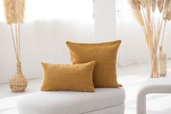 Zwei gelbe Kissen auf hellem Möbelstück, mit strukturierter Oberfläche und dekorativen Pflanzen im Hintergrund.