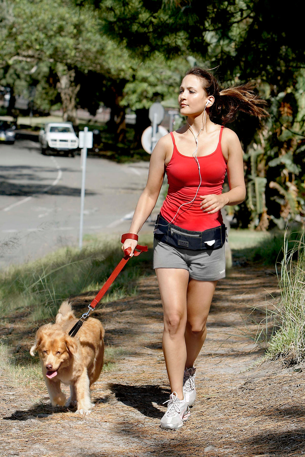 Junge Frau im roten Top mit Hund am Weg, trägt Kopfhörer und Umhängetasche, Bäume und Straße im Hintergrund.