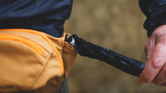 Person mit orangefarbener Bauchtasche, aus der ein schwarzer Regenschirm gezogen wird, unscharfer Hintergrund.