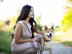 Frau mit langen, dunklen Haaren sitzt lächelnd auf dem Boden mit einem leinen Hund im beigefarbenen Outfit.