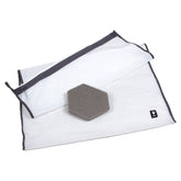 Weißer Wäschebeutel mit Reißverschluss, dunklem Rand und schwarzem Etikett, neben grauem sechseckigem Schwamm.