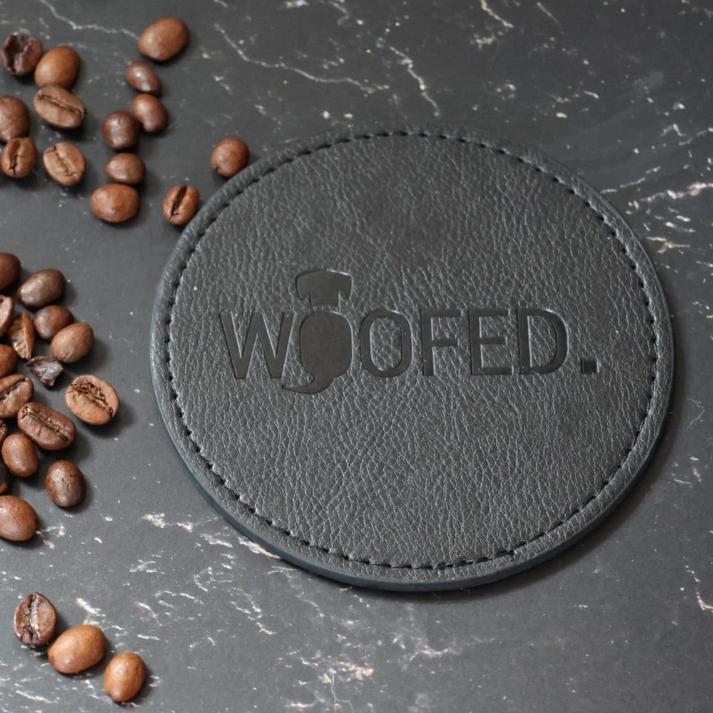 Runder schwarzer Leder-Getränkeuntersetzer mit „WOOFED“-Prägung, umgeben von Kaffeebohnen auf dunkler Oberfläche.