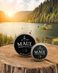 MAUL Bio Pfotenbalsam-Dosen inmitten einer malerischen Landschaft mit See und Wäldern.