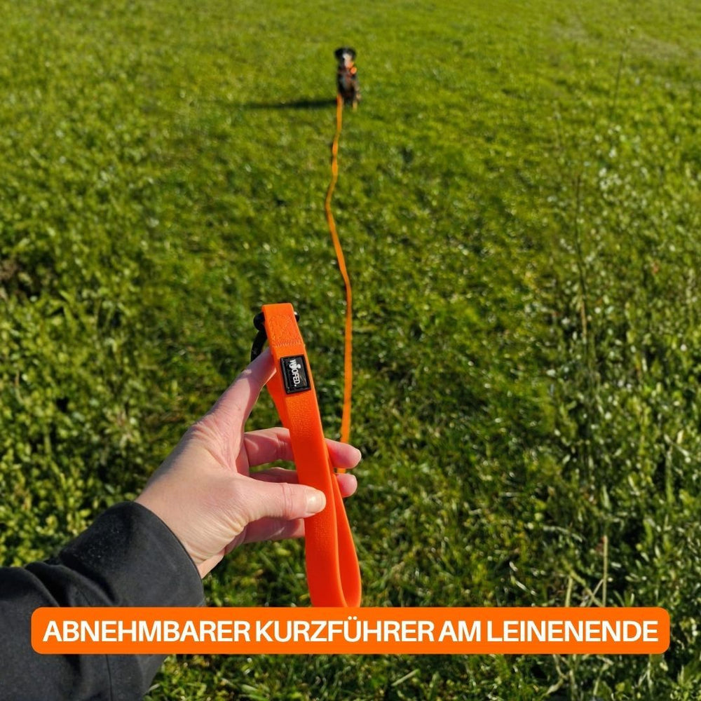 Hand hält orangefarbene WOOFLEX® Hundeleine mit abnehmbarem Kurzführer vor grünem Wiesnhintergrund.