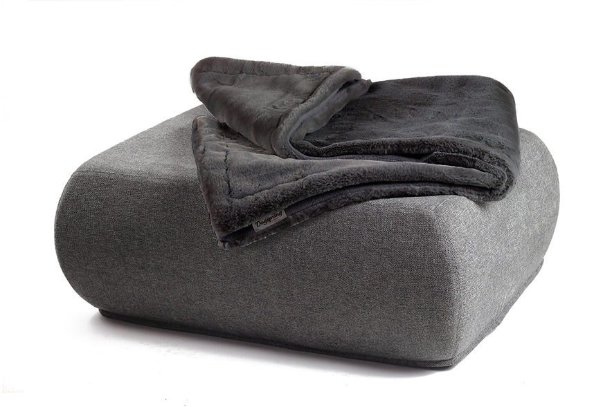 Graue Wohndecke aus Webpelz auf einem modernen Pouf mit abgerundeten Kanten vor weißem Hintergrund.