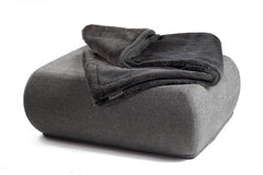 Graue Wohndecke aus Webpelz auf einem modernen Pouf mit abgerundeten Kanten vor weißem Hintergrund.