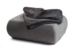 Graue Wohndecke aus Webpelz auf einem modernen Pouf mit abgerundeten Kanten vor weißem Hintergrund.