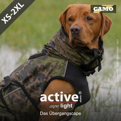 Hund im camouflaged Übergangsparka "active cape light" auf Wiese, umgeben von Gras und Wasser.