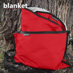 Rote Decke mit grauem Innenfutter, zusammengefaltet und von schwarzer Umrandung gehalten, vor einem Baumstamm.