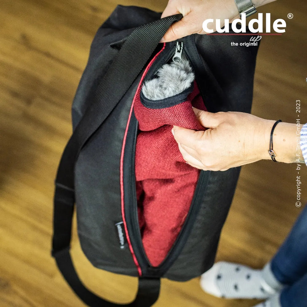 Schwarze Tasche mit rotem Innenfutter für den Cuddle.up® Hundeliegeplatz auf Holzoberfläche geöffnet.