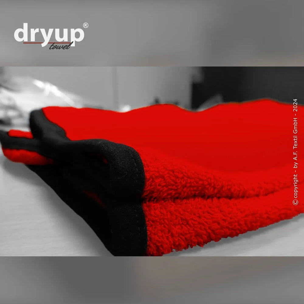 Rotes Dryup® Handtuch mit schwarzem Rand, gefaltet auf einer Oberfläche, Markenname oben links sichtbar.