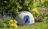 Rundes, graues Insektenhaus mit blauer Öffnung im bunten Blumengarten mit Tulpen und Lavendel umgeben.