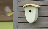 Nistkasten für Vögel an grauer Wand, mit runder Öffnung und abgerundetem Dach, während ein Vogel im Hintergrund fliegt.