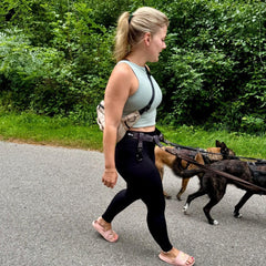 Frau mit blonden Haaren geht mit mehreren Hunden an verstellbarem Gassigürtel in der Natur spazieren.
