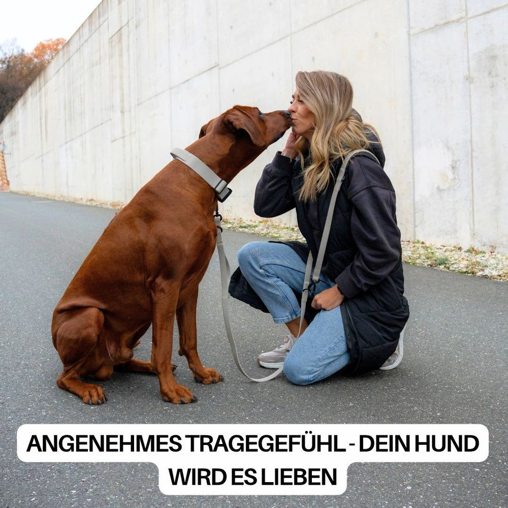 Frau mit dunkler Jacke und heller Hose kniet und lächelt, während ihr brauner Hund sie zärtlich küsst.