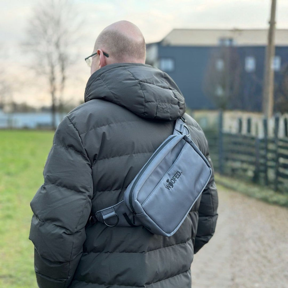 Mann mit grauer Gassitasche in der Natur, trägt sie als Bauchtasche und Schultertasche in grüner Umgebung.