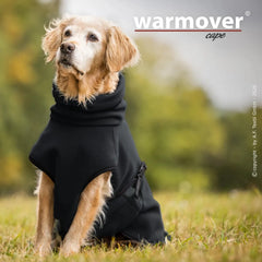 Hund im Warmover© Cape sitzt bequem auf einer Wiese, umgeben von verschwommenen Bäumen.