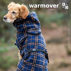 Hund im blauen, karierten Warmover-Cape mit hohem Kragen und Bauchgurt vor unscharfem Naturhintergrund.