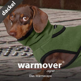 Dackel trägt grünes warmover© Wärmecape aus weichem Material mit hohem Kragen auf gemustertem Boden.