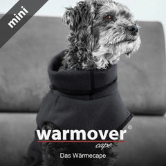 Kleiner Hund mit schwarzem Fleece-Wärmecape auf grauer Couch, aufmerksam zur Seite schauend.