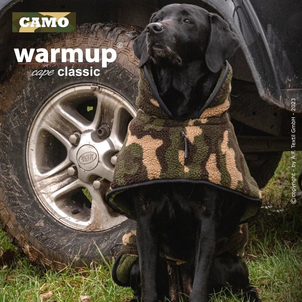 Schwarzer Hund mit camouflagemustertem Wende-Cape sitzt neben einem Auto in natürlicher Umgebung.