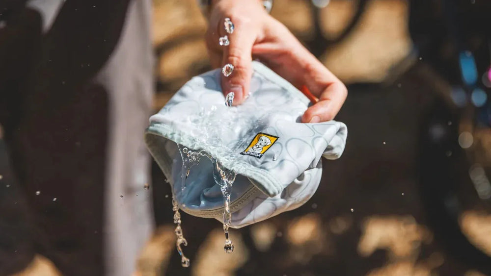 Graues Swamp Cooler™ Halstuch in der Hand mit spritzendem Wasser, Fahrräder im unscharfen Hintergrund.