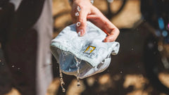 Graues Swamp Cooler™ Halstuch in der Hand mit spritzendem Wasser, Fahrräder im unscharfen Hintergrund.