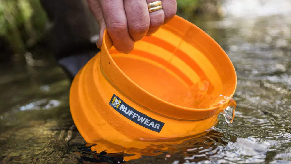 Eine Hand hält den orangefarbenen Bivy™ Faltbaren Hundenapf von Ruffwear, während Wasser herausfließt.