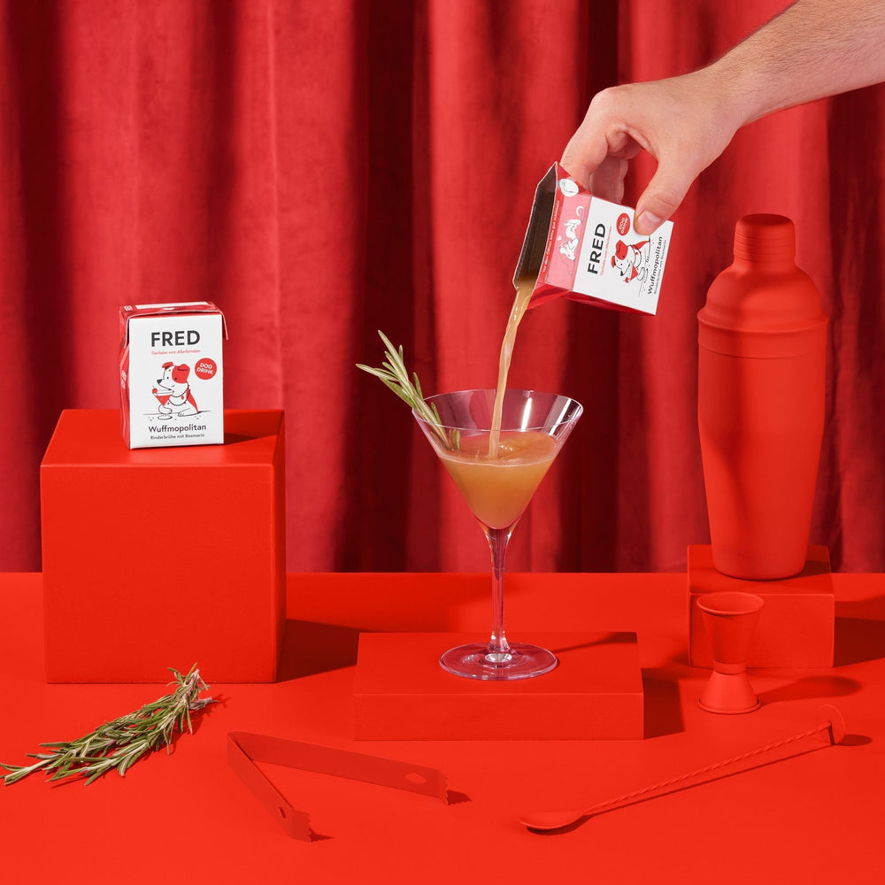 FRED Dog Drink "Wuffmopolitan" 2er Set für Hunde, stilvoll präsentiert mit roter Shaker-Flasche und Rosmarin.