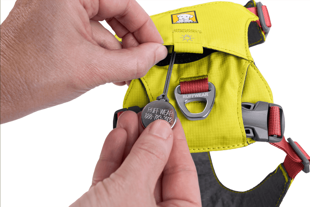 Person hält gelbes Hi Light™ Hundegeschirr von Ruffwear mit geöffnetem Verschluss und ID-Tag vor gelbem Hintergrund.