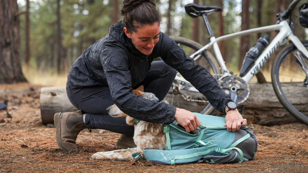 Person mit schwarzer Jacke öffnet Hitch Hiker™ Hunde Rucksacktrage im Wald, während Hund davor sitzt.