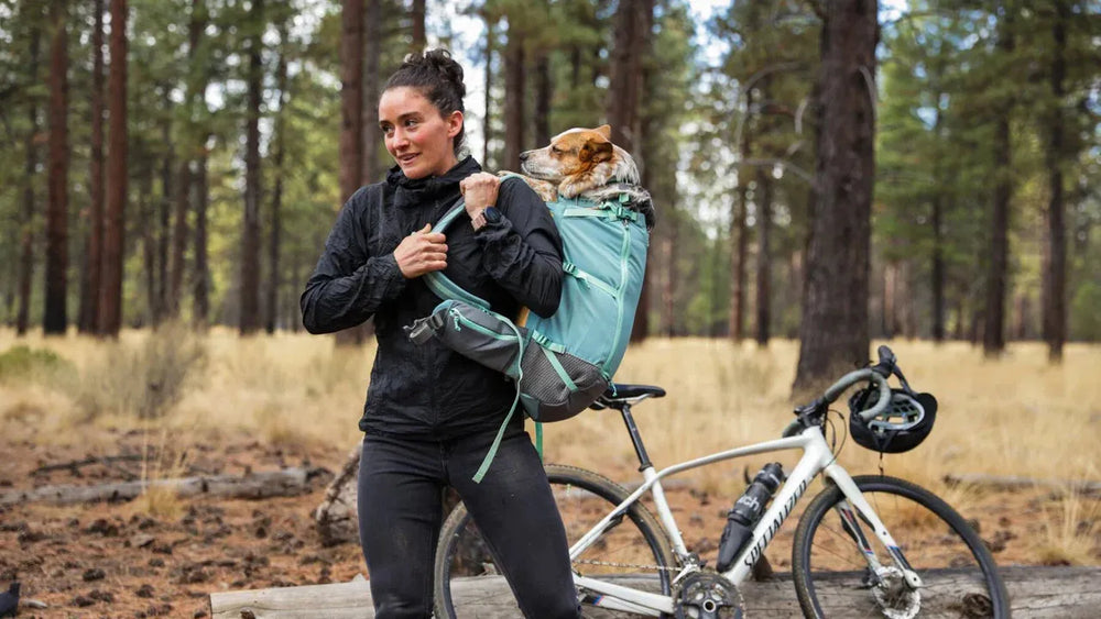 Frau mit Hitch Hiker™ Hunde Rucksacktrage im Wald, lächelt während ihr Hund herausblickt. Fahrrad in der Nähe.