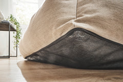 Beige CLASSIC Hundebett SMOOTH Unterseite mit Schutznetz auf Holzboden, Hintergrund unscharf mit Tisch und Pflanzen.