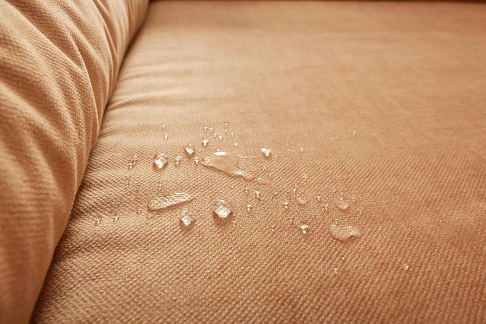 "Beigefarbenes ORTHO Hundebett GLAM mit gepolsterter, wasserabweisender Oberfläche und sichtbaren Wassertropfen."