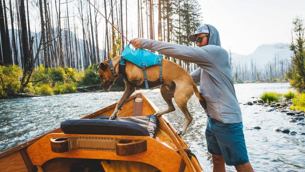 Mann hebt Hund in Confluence™ Schwimmweste auf Boot in natürlicher Landschaft am Fluss an. Freundliche Atmosphäre.