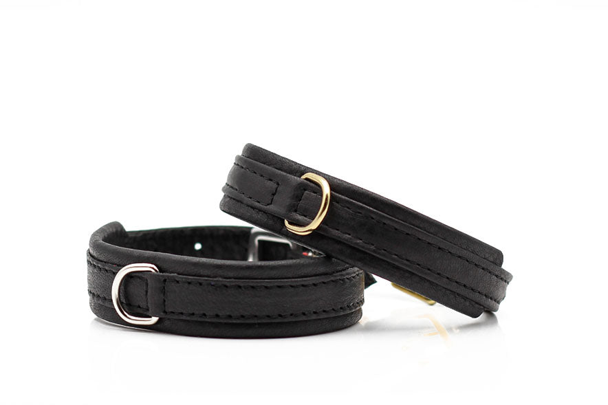 Zwei schlichte schwarze Lederarmbänder mit goldfarbener Öse, elegant und minimalistisch, nebeneinander angeordnet.