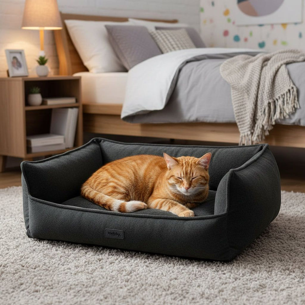 "Gemütliches, dunkles Katzenbett 'NILA' mit entspannter orangefarbener Katze im Schlafzimmer."