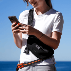 Person mit DOOG Neosport Hip Belt in Schwarz vor blauem Himmel und Gewässer, hält ein Smartphone in der Hand.