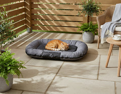 Gemütliches Katzenbett "RENO" auf einer Terrasse, umgeben von Pflanzen und einem geflochtenen Stuhl.