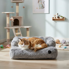 Gemütliche graue Sofamatte "NURU" mit entspannter orange-weißer Katze, im Hintergrund Kratzbaum und Spielzeuge.