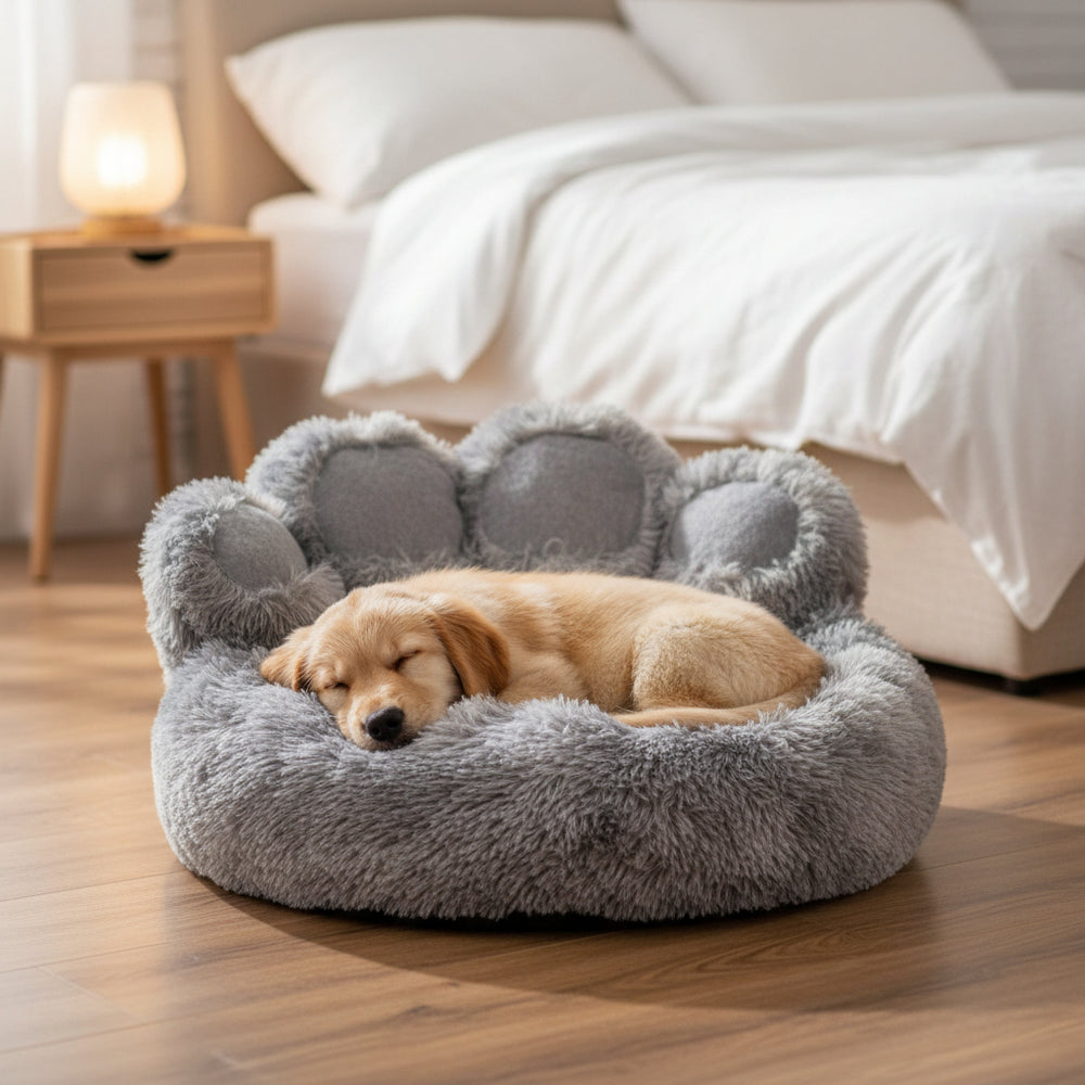 Schlafender Welpe in grauem, runden Hundebett in Pfotenform mit weichen Rändern auf einem gemütlichen Hintergrund.